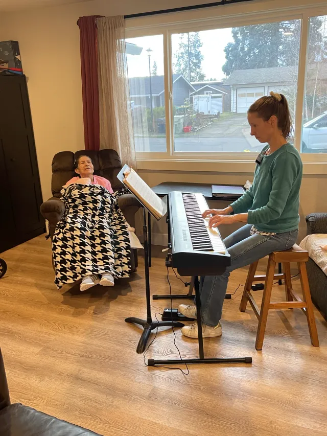 A caregiver plays an electric piano in the living room while a resident listens from a recliner.